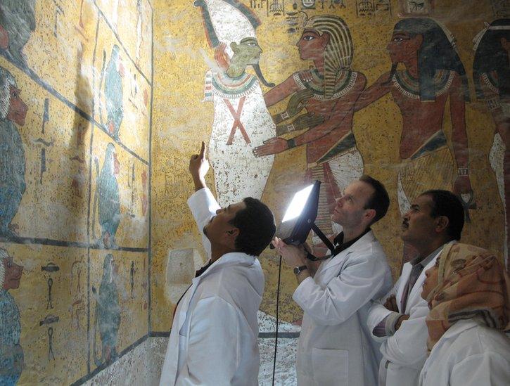 3 conservationists in white lab-coats hold up a light to inspect wall-paintings