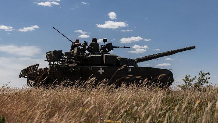 A large Ukrainian army tank in a field with long beige grass or crops,  pictured against a blue sky. Two soldier outlines visible sitting on top of vehicle.