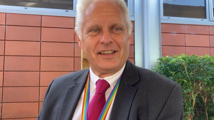 Head teacher Neil Morris speaks outside a school building. He is wearing a grey suit, pink tie and rainbow lanyard