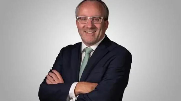 Richard Corrigan is wearing a dark blue blazer and green tie. He has glasses and is in front of a white background. 