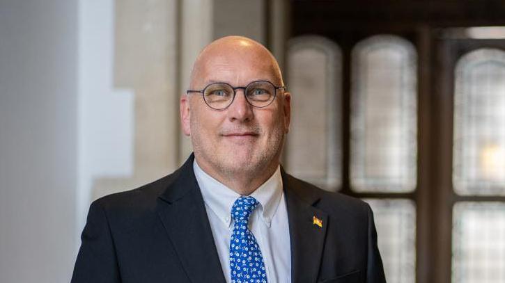 Nick Adams-King smiling for the camera inside a building. He is wearing glasses and a dark suit with a light blue white-dotted tie