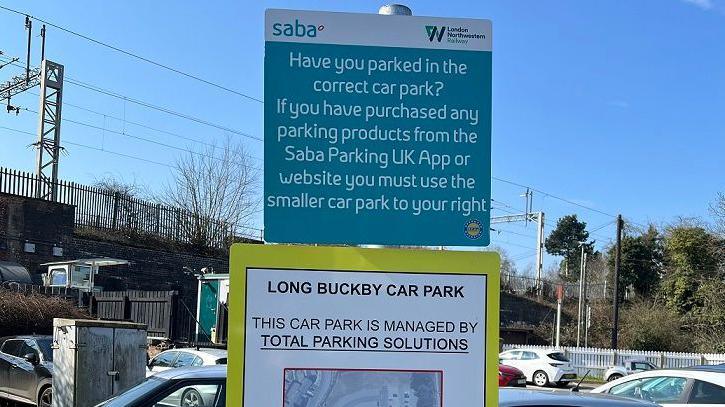 Sign in a car park in white writing with blue background, which states that drivers who have paid Saba must park in the smaller car park. It includes a diagram.  There are cars parked behind the sign and a railway line is visible to the left.