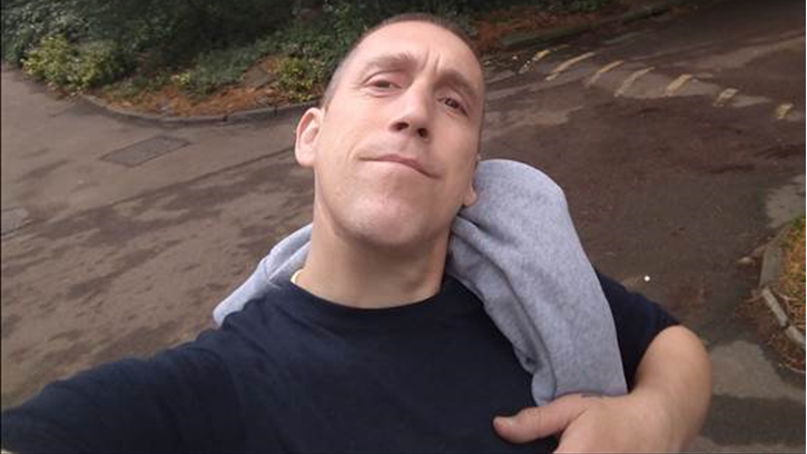Matthew Cainey wearing a dark top with a grey hoodie over one shoulder. He has the camera up high taking a selfie of himself, smiling at the camera.