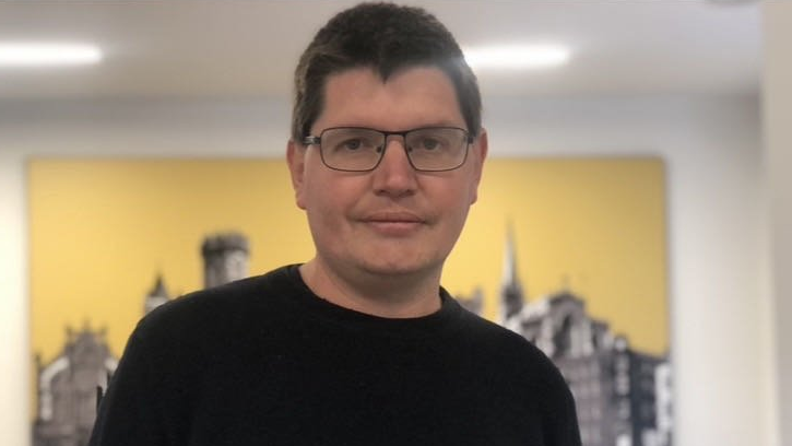 Shows a man with dark hair and glasses and a black round-neck jumper standing in front of a wall with a poster of buildings on it.