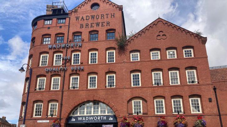 The old Wadworth brewery, a large red brick building. There is a large arch in part of it. It has multiple white-framed windows.