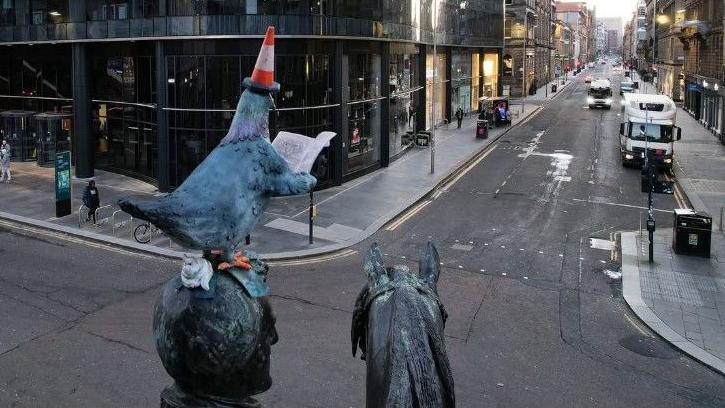 A view from behind of a statue, showing a pigeon with a traffic cone sitting on top of another statue. 