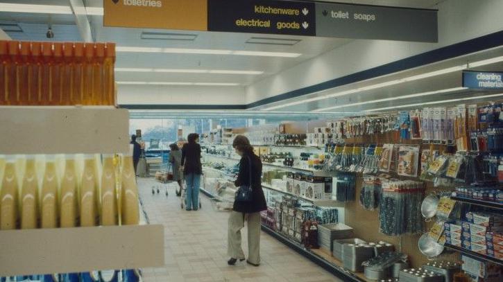 An interior shot of Sainsbury's, Cambridge, in about 1975. It shows women in an aisle and on their right are piles of kitchenware and electrical goods