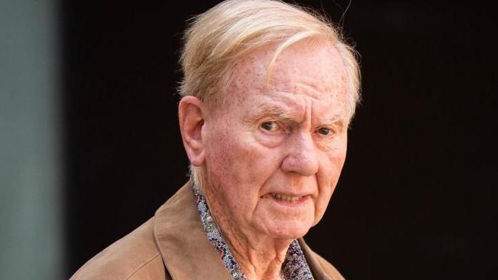 An older man in a pale brown jacket and paisley shirt looks toward the camera with a serious expression.
