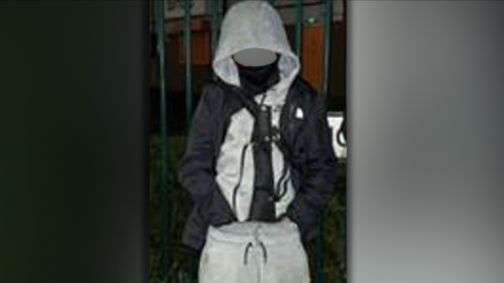 A boy in a grey tracksuit, with a machete in a black covering tucked into his trousers. His face is blurred and he is standing outside in the dark in front of a fence