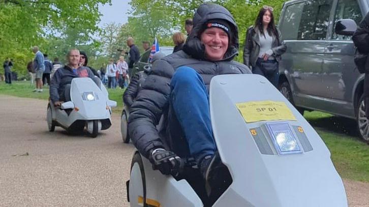 Bryan Griffin is smiling as he rides in his C5 vehicle. He is wearing a padded anorak, blue trousers and gloves. There are other C5s behind him and a woman is walking past some parked cars next to the pathway