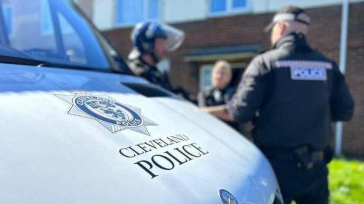 The front bonnet of a white van bearing the title and the logo of Cleveland Police, with an out of focus view of three police officers, one with his back to the image, another on side view wearing a helmet and a visor and a third facing