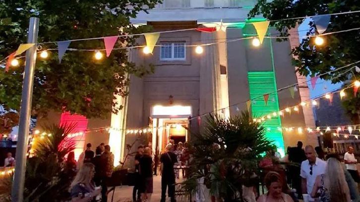 A crowd of people gather outside the St James hall in Guernsey during an evening event with bunting and lights decorating the area in front of the building.