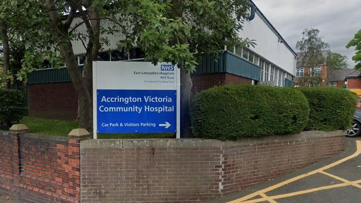 A sign outside the Accrington Victoria Community Hospital. It has directions to the car park and visitor parking. The sign is next to trees and bushes and surrounded by a brick wall.