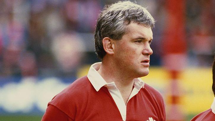 A man with grey-brown hair wearing a red Welsh rugby jersey. He has a serious expression on his face as he looks off to the side. There's a blurry background of a rugby stadium behind him.