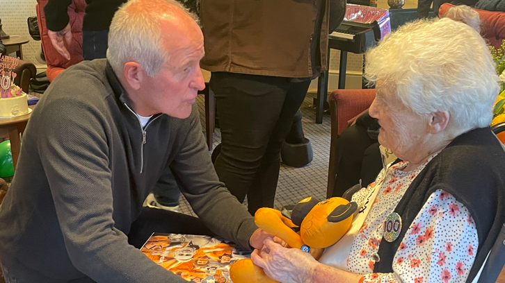 Steve Bull is kneeling and facing Joan in her chair and holding her hands while he talks to her. Joan is smiling at him and is holding some Wolves memorabilia and is wearing a 100 birthday badge.
