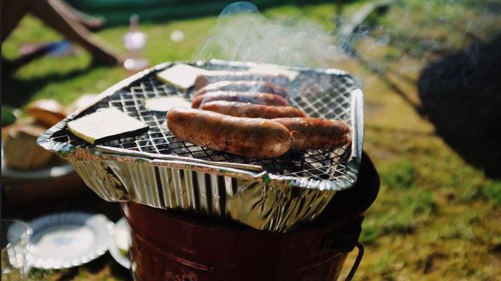 The picture shows a disposable barbeque placed on a bucket. It has a silver casing, and silver wire goes across the top in a criss-cross pattern. Sausages are cooking and there is smoke coming from them. There are various bowls, plates and cups in the background.
