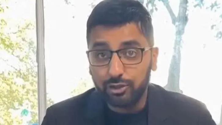 MP Adnan Hussain in front of a window showing trees in the background with black hair and black beard wearing glasses and a dark grey suit jacket and black t-shirt.