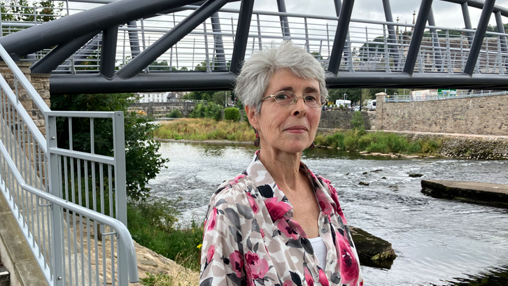 Ms Dales is wearing a pick and grey shirt and is standing in front of a river, with a bridge overhead.