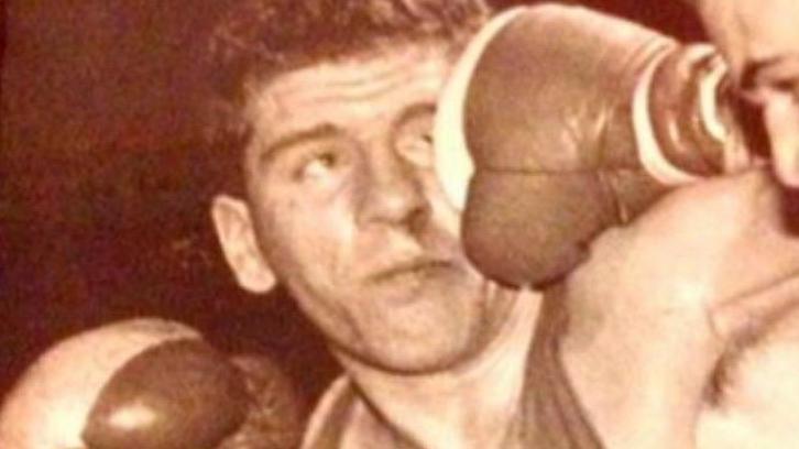 A sepia photo of a young man wearing a singlet and boxing gloves. He is holding one fist up near his face and his other arm is outstretched. Someone out of shot is punching him in the face.