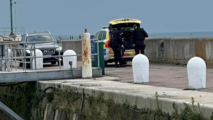 a police vehicle parked on Ramsgate Harbour with boot open while two officers in black uniforms, carrying what appears to be guns, stand with their backs to the camera