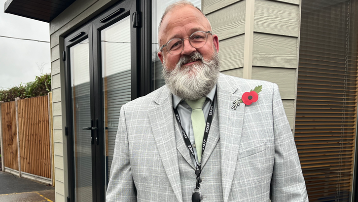 Glyn Hambling stands in front of a pale green cabin structure. He is wearing a light grey checked suit with a red poppy attached to it. He has grey hair and a grey beard.