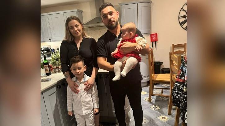 Sophia Kelemen is held by her dad, who is standing next to her mum and her big brother. They are in the kitchen and dining area of a house. Sophia's mum and dad are smiling and looking at the camera.