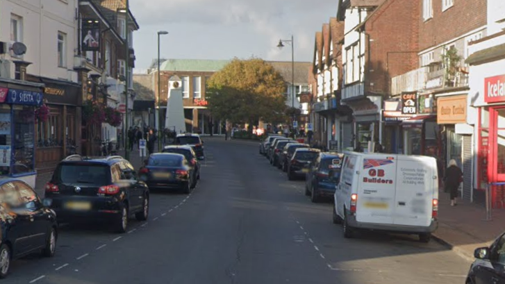 A Google Street image of a road with vehicles parked either side and shops either side.