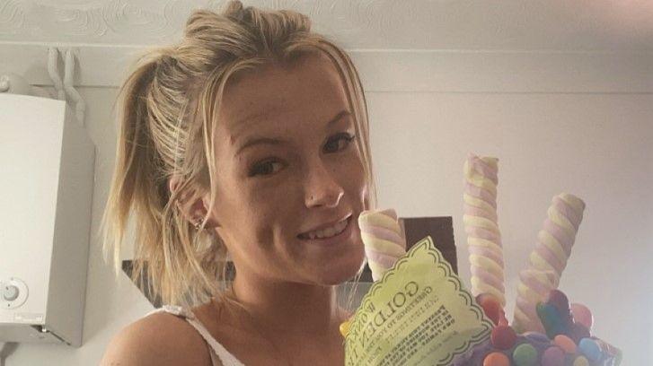 A young blonde woman with her hair tied in a ponytail. She is smiling at the camera and holding a cake covered in Marshmallows, chocolate sweets and a piece of yellow paper which reads "Golden Ticket".