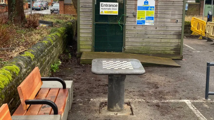 A chess table in a car park front of a wooden cycle storage shed