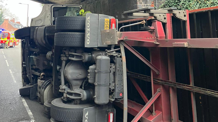 The lorry lies on its side under the bridge with emergency vehicles parked nearby