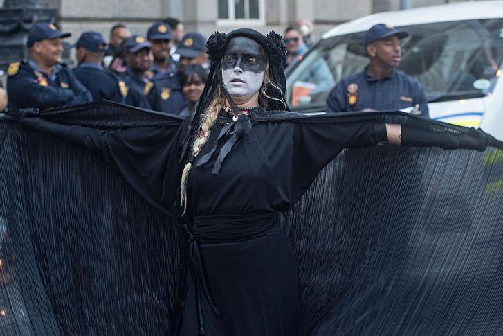 Woman wearing all black and black and white face paint spreads open her cape as she protests with police behind her.