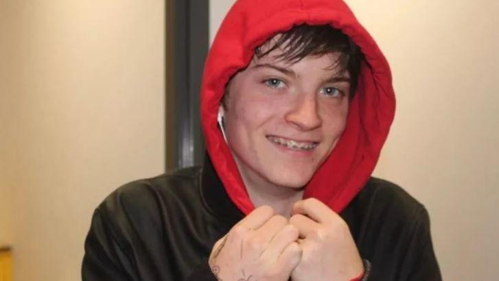 Daniel Davies smiling at the camera. He has braces on and white earphones in. He is wearing a black jacket and a red jumper with the hood pulled up. His hands are gently pulling at the front of the hood.