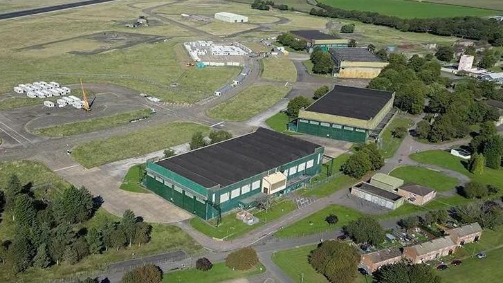 An aerial view of the former RAF Scampton site: two defunct military buildings stand in the middle of green open spaces with pine trees on the left and right of the picture. On the top right there are small white cabins nestled in two clusters.