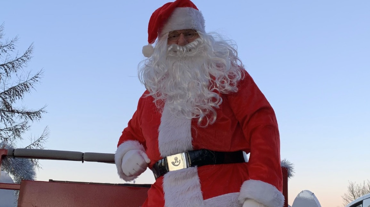 A man dressed as Santa with a large white beard and red hat with a white bobble on the end is leaning on the railing of a sleigh.