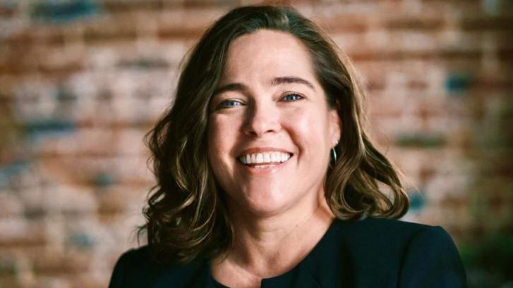 Dr Melissa McCullough is smiling widely at the camera. She has shoulder-length hair which is brown with caramel highlights. She is wearing a black top. Behind her is an exposed brick wall.