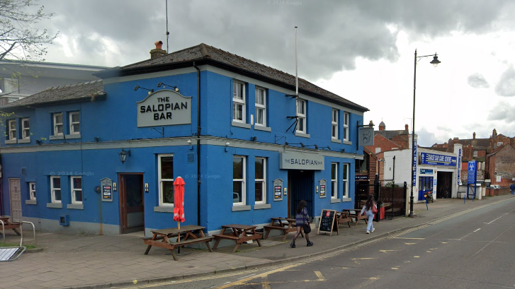 The Salopian bar is painted blue, and has wooden benches outside it. It it located next to a road. 