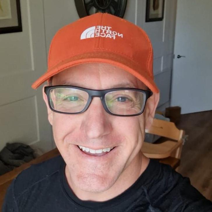 Ed Stroud is smiling and is wearing black, rimmed glasses and an orange The North Face cap on his head.