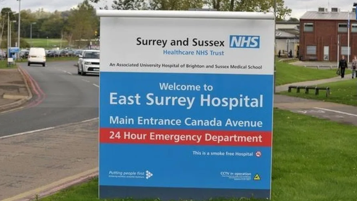 The entrance sign to East Surrey Hospital, with white lettering on a blue background, red brick buildings, roads, grassed areas and parked cars in the background.