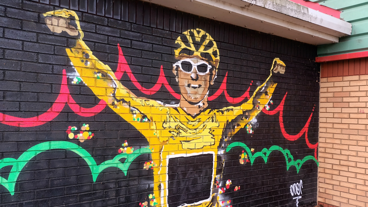 A colourful mural of Geraint Thomas wearing the yellow cycling jersey and helmet and white cycling glasses. Painted on a black background on a brick wall, with red and green swirls behind him.