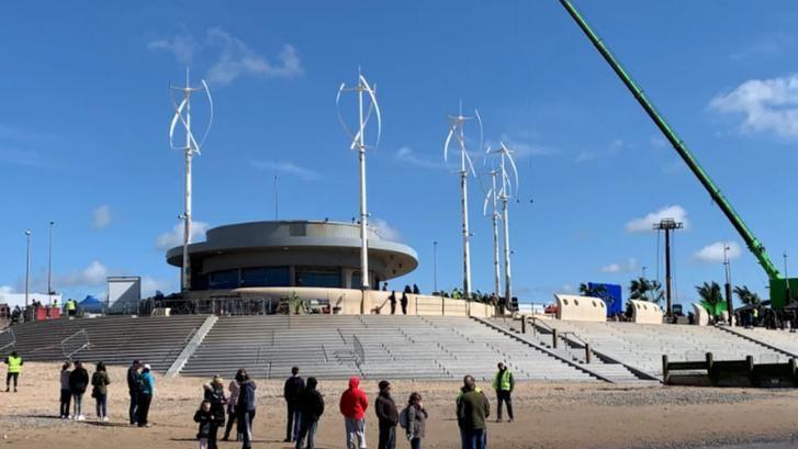 People stand on a beach below some stairs. A green crane is in the background. 