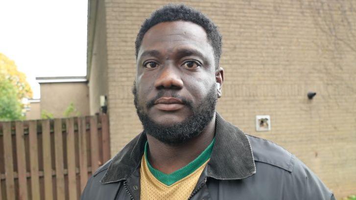 A man stands in front of a red brick wall and wooden slat fence. He is looking a the camera and has short hair and a trimmed beard. He is wearing a grey jacket and a yellow and green football shirt