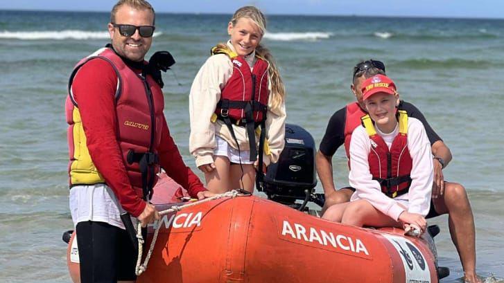 Two girls are wearing life jackets and are aboard a blow-up boat. The jackets are red. Annie is wearing a red hat. There are two adults next to them keeping the boat steady.