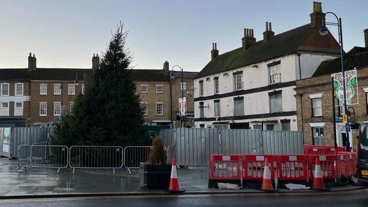 St Neots Market Square, with a Christmas tree, metal barriers, red barrier, red cones, Christmas lights and buildings around it, including the boarded-up Falcon Hotel.