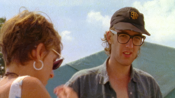 Robin as a young man looking to camera, with black glasses and a dark baseball cap and shirt with a woman in sunglasses visible from the back of her head standing next to him. There is a green tent behind them.
