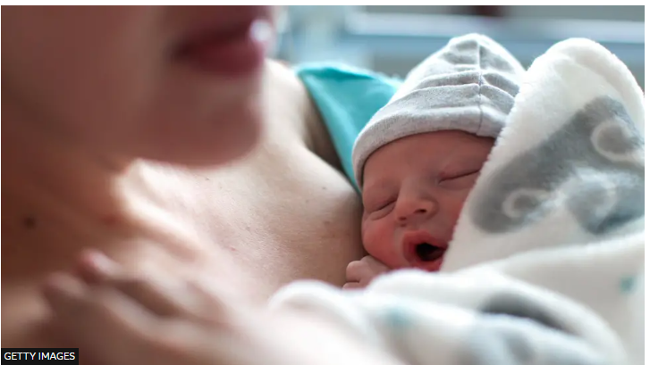 Close-up of a mum holding her sleeping newborn baby on her chest. Baby is wearing a pale coloured hat and is wrapped in a blanket.