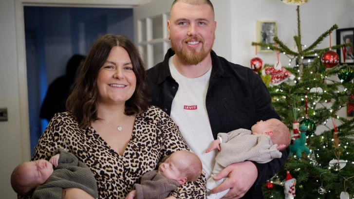 Stacey and Grant standing together, smiling to camera, with mum holding two triplets, and dad the other one; all are fast asleep. There's a Christmas tree in the room in the background  