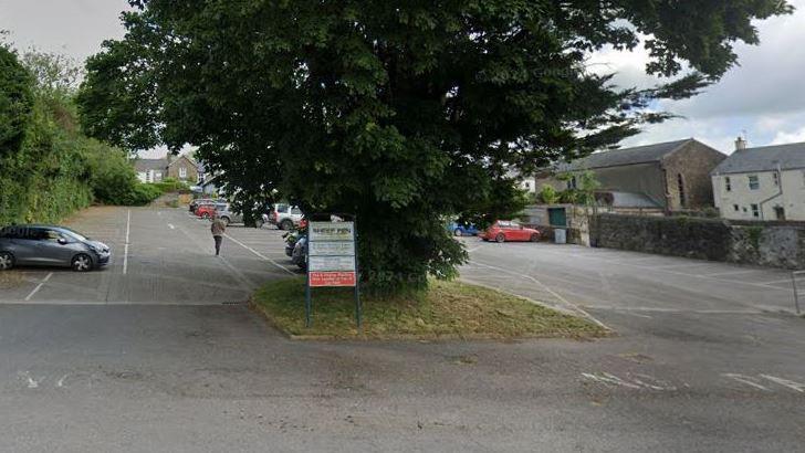 A car park with a tree and houses on the edge and some shrubbery with a sign and a car on left and some more in the distance.