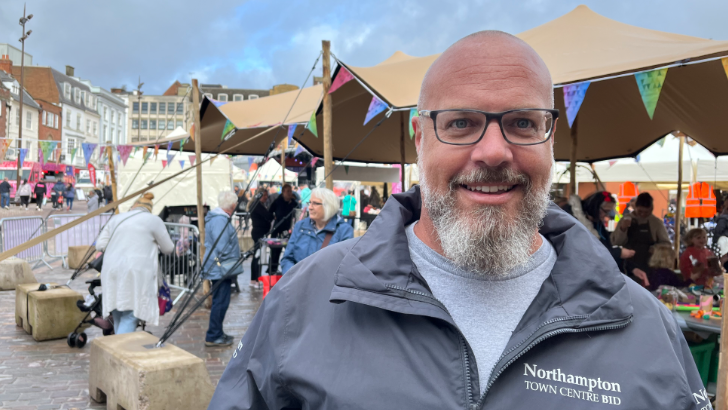 Man with grey beard, glasses and bald head smiling at camera, wearing a light grey tshirt and darker grey zip-up jacket with "Northampton Town Centre Bid" in white lettering to one side of the chest. Behind him and obscured by him is a stall covered with an awning and surrounded with coloured bunting and various people standing around and buildings around the market square.