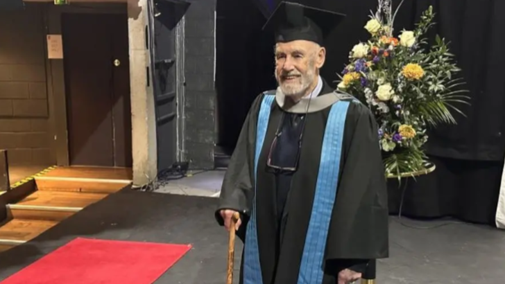 A man walking across a graduation stage. He is wearing a gown with a blue tab and a black cap. He holds a walking stick. There is a large bunch of flowers in the background.