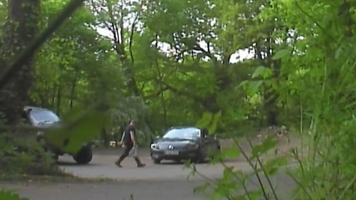 Still image of surveillance footage with a some foliage in the foreground and showing a man wearing black walking to a black car with a supermarket carrier bag in his hand. He is walking on tarmac and behind him and car is a dense woodland